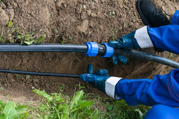 worker's hands hold a black pipe over a dug hole in the ground