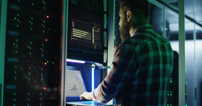 Adult bearded man setting server hardware and speaking on smartphone in data center corridor