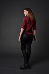 full length portrait of brunette girl wearing  red shirt and leather pants. standing pose, facing away from the camera, on grey studio background.