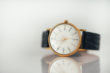 Vintage golden watch laying on white glossy surface with reflection. Copy space. Black leather strap.  