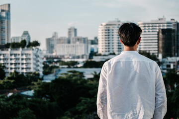Young asian man sitting on the edge of the roof and looking far away at the city