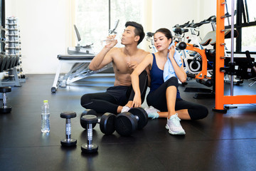 Fototapeta na wymiar Attractive young sports people are holding bottle of water, talking and smiling while resting in gym