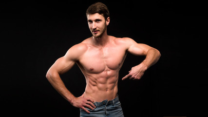 Fototapeta na wymiar Handsome young muscular man shirtless wearing jeans, on dark background in studio shot. Showing his ABS by finger.