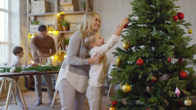 Family preparing home for celebration: happy mother and daughter decorating Christmas tree together while father packing gift boxes with little son