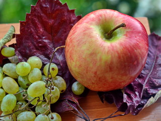 sprig of green grapes and a red ripe Apple on the autumn leaves of a vine on a wooden table