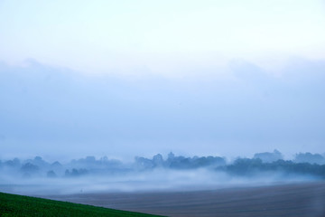 British countryside looking across a sweeping field to an old english village covered in white morning mist, the trees and the buildings are above the mist and the church tower can be seen silhouetted