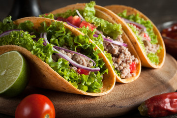 Photo of Mexican tacos with ground beef, onion, tomatoes, chili, red sauce, lettuce and lime on wooden background. Spicy and fast food concept.