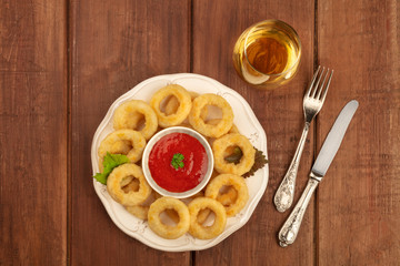 An overhead photo of squid rings with a red sauce, a glass of white wine, and vintage cutlery, shot from above on a dark rustic wooden background with a place for text