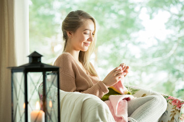 Young woman sitting home in a chair by the window wearing knitted warm sweater, knitting with needles. Cozy room decorated with lanterns and candles. Scenic winter view of pine trees in snow in window