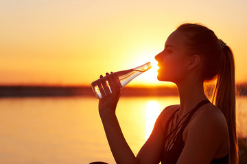 Silhouette woman drinking water from bottle after run or yoga on the beach. Fitness female profile at sunset, concept of sport and relaxation