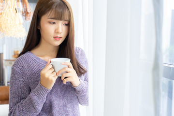 Asian woman holding a cup of coffee in her hands on the light background from window.She dress sweater violet colour.Morning coffee  time.