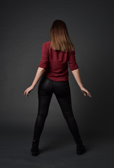 full length portrait of brunette girl wearing  red shirt and leather pants. standing pose, facing away from the camera, on grey studio background.