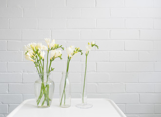 Three glass vases with white freesia flowers on small table against painted brick wall with copy space