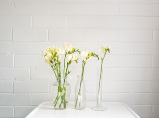 Three glass vases with white freesia flowers on small table against painted brick wall