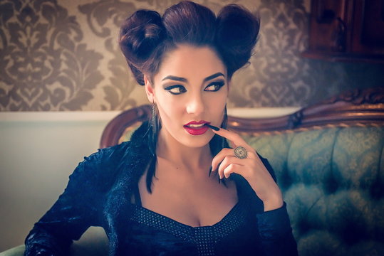 Sexy Caucasian model looking like a vampire with a black stiletto nail between the teeth, sculpting make-up and cocktail dress with big cleveage. Maleficent lookalike retro concept