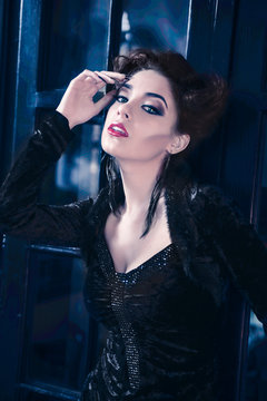 Sexy Caucasian model like a vampire looking straight and standing against a window door, with big bright eyes, sculpting make-up and cocktail dress with big cleveage. Maleficent lookalike concept