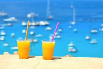 bright orange cold refreshing smoothie drink in a plastic cup on stone wall with turquoise blue sea with boats and beach in background on a hot sunny day. Travel concept
