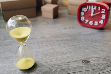 Hourglass as time passing concept with red clock and cardboard box on wooden background.