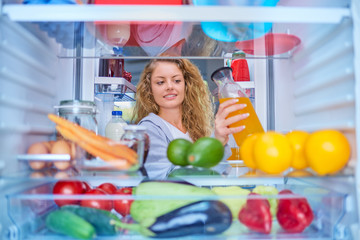 Woman standing in front of fridge full of groceries and taking juice. Picture taken from inside of...