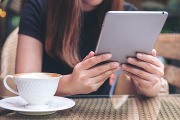 Closeup image of a woman holding and using tablet pc with coffee cup on table in cafe