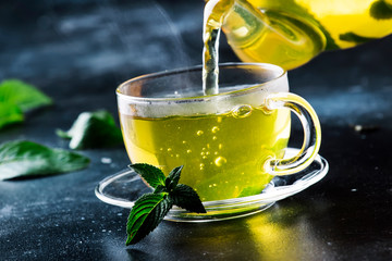 Hot chinese green tea with mint, with splash pouring from the kettle into the cup, steam rises, dark background, selective focus