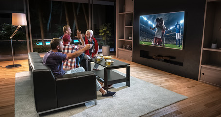 Group of fans are watching a soccer moment on the TV and celebrating a goal, sitting on the couch in the living room.