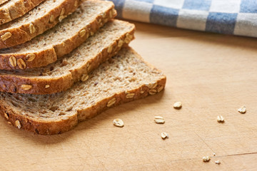 Close-up of slices of whole grain bread on a wooden cutting board with bran and a blue and white checkered cloth in the background