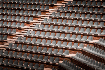 Free unclaimed seats in multiple rows. Sunset photo in empty public arena and concert amphitheatre. Red brick and white travertine structure, gray plastic chairs. 