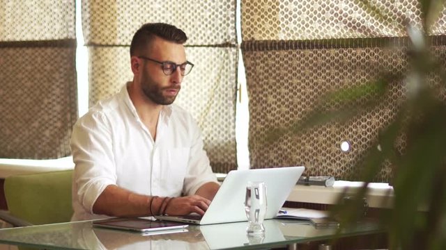 Bearded young man working in office, laptop, carefully looking through glasses, smiling. Freelancer, roman blinds
