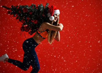 Santa hat Christmas sporting woman holding  xmas tree on her shoulders. Winner energy he red background. it's snowing