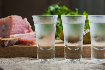 Friendly booze - three glasses of vodka on the table on the background of snacks - lard, garlic, parsley, bread, cucumber