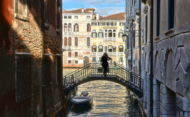 Picturesque scene Silhouette of young woman on small old bridge between olden buildings in narrow water canal and facade of historical buildings and Canal Grande in background. Venice Italy