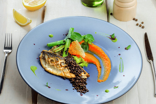 Plate with delicious grilled seabass fish fillet steak and vegetables on a white wooden table. Healthy gourmet food.
