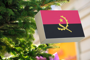 Angola flag printed on a Christmas gift box. Printed present box decorations on a Xmas tree branch on a street.