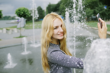 Portrait of beautiful student teenager girl holding up a smartphone taking selfies photos, networking using technology, outdoors park. Adolescent with books near water fountain, lifestyle exterior.