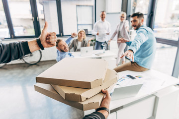 Obraz premium cropped shot of man holding pizza boxes and showing thumb up to colleagues in office