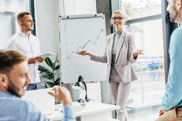 smiling young businesswoman standing near whiteboard and looking at male colleagues in office