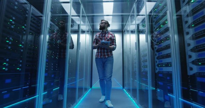 Wide shot of casual man with tablet walking among server racks in data center room looking at hardware