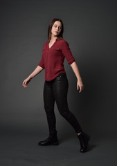 full length portrait of brunette girl wearing  red shirt and leather pants. standing pose, on grey studio background.