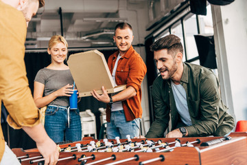 group of young casual business people playing table football with pizza at office and having fun...