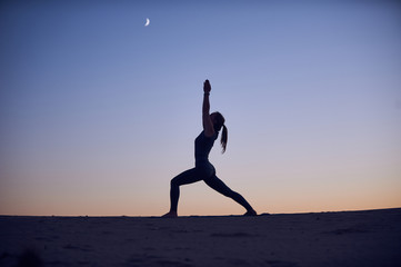 Beautiful young woman practices yoga asana Virabhadrasana 1 - warrior pose 1 in the desert at night