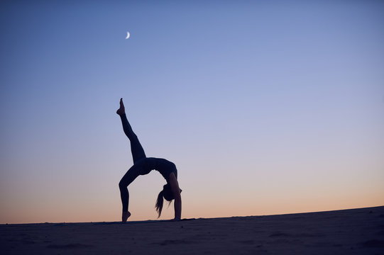Beautiful young woman practices backbend yoga asana Eka Pada Urdhva Dhanurasana - One Leg Upward facing bow pose in the desert at night