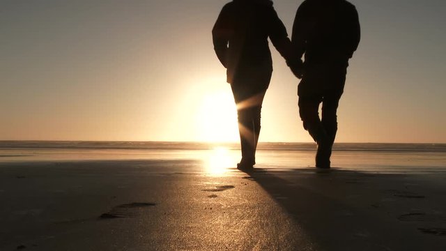 Silhouetted beach couple walk out on sandy coast to watch beautiful ocean sunset together.