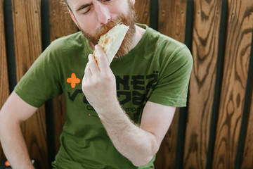 Street food. hipster man with a beard on the street on the bench eating doner kebab. wooden background 