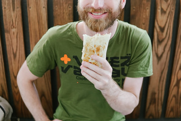 Street food. hipster man with a beard on the street on the bench eating doner kebab. wooden background 