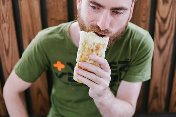Street food. hipster man with a beard on the street on the bench eating doner kebab. wooden background 
