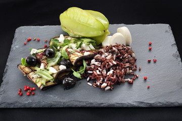 Sałatka z grillowanym bakłażanem. Grillowany bakłażan z czarnymi oliwkami, serem feta i zielonymi warzywami podany z dzikim ryżem na czarnym kamiennym talerzu.