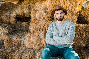 Naklejka na ściany i meble confident male farmer in sweater and straw hat sitting with crossed arms on hay stacks at ranch