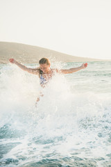 Young beautiful blond woman tourist in swimsuit standing and enjoying splash of wavy waters of Mediterranean sea in Prasonisi cape, Rhodes, Greece on summer day