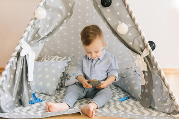 Cute one year old baby playing with smartphones a tent, wigwam. Shadow depth of field, natural light. The interior of the children's room is grey, blue and white